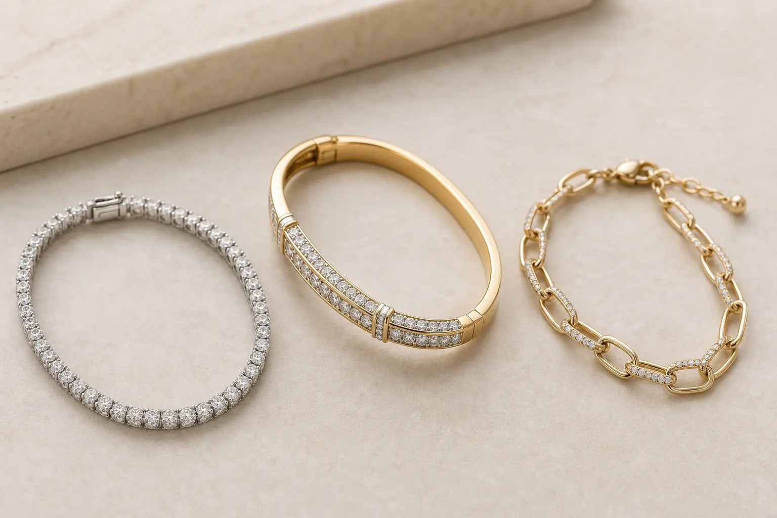 Three diamond bracelets on a neutral surface: a tennis bracelet, a bangle, and a chain bracelet.